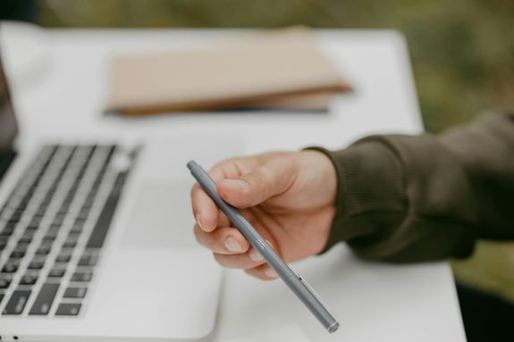 Close-up of a person holding a ballpoint pen near a laptop, symbolizing work or study.