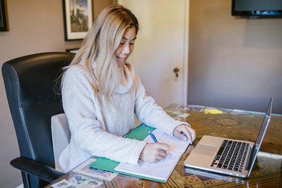 Asian woman working from home, signing documents at a desk with a laptop.