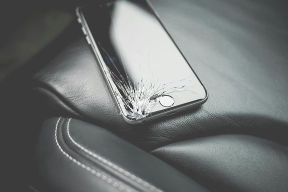 Close-up of a broken smartphone with a cracked screen on a leather car seat.