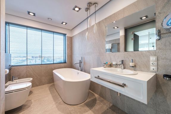 White bathtub and bidet placed near sink with hygienic supplies and mirror in stylish light bathroom with jalousie on window