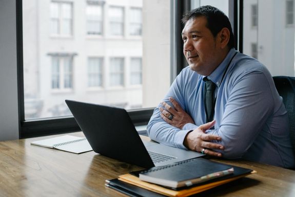 Adult successful ethnic male boss wearing shirt and tie sitting with hands crossed at workplace with documents and netbook