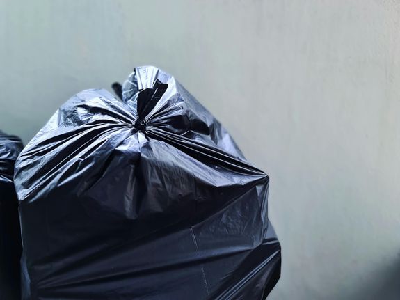 A close-up photograph of a black plastic garbage bag against a plain concrete wall, highlighting waste management.