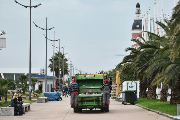 A vibrant urban scene featuring a garbage truck on a street lined with palm trees and people enjoying the promenade.