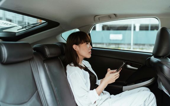 Side view of focused young Asian female wearing casual white clothes and using modern smartphone while riding at car backseat